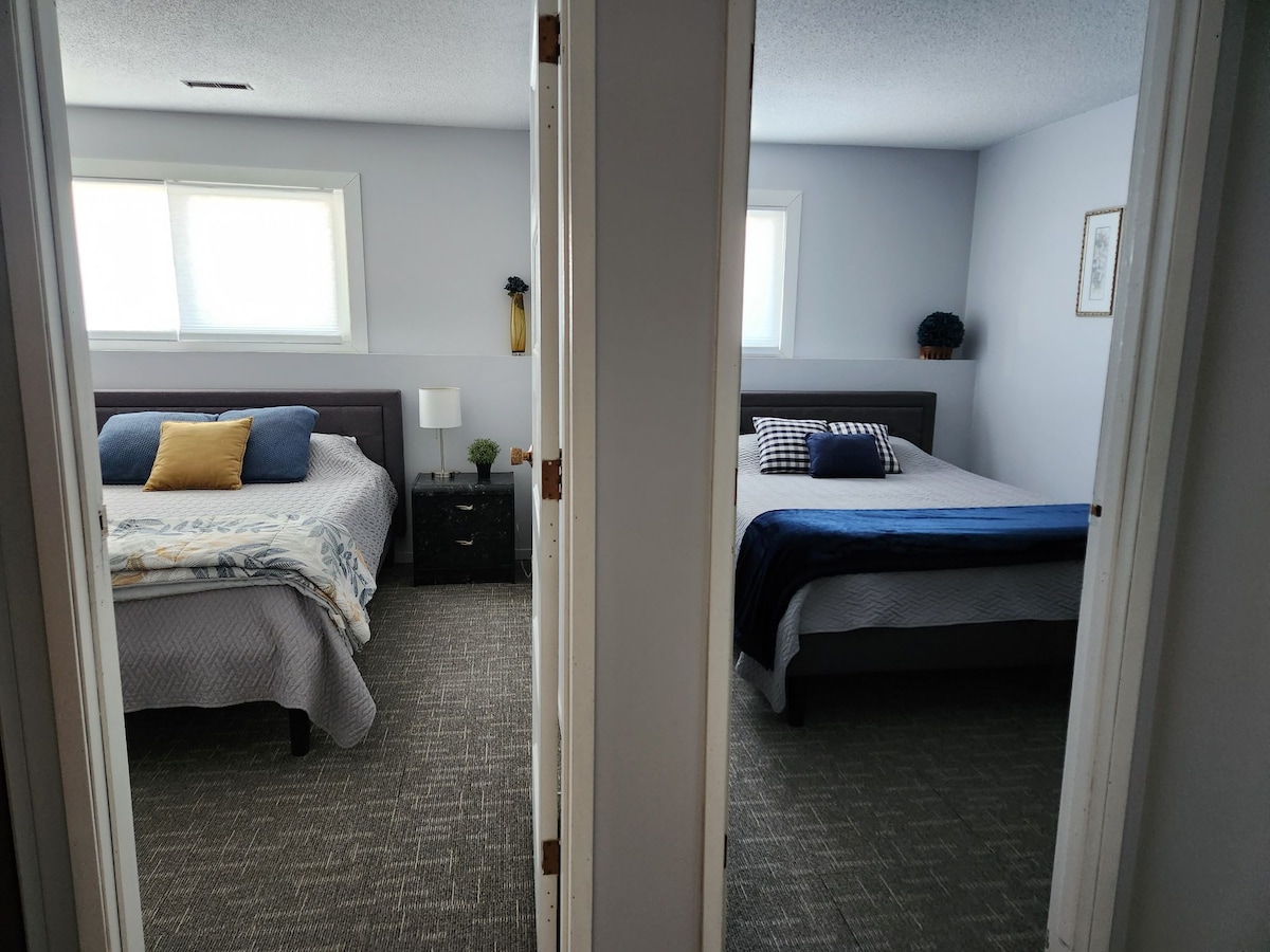 Two inviting bedrooms are seen through an open doorway. Each bedroom features a queen bed with bedding in calming tones. Nightstands are placed beside the beds, and both rooms include windows that allow natural light to enter, enhancing the serene atmosphere.