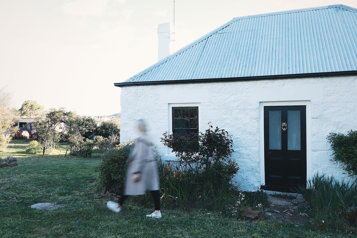 A rustic cottage with a light-colored stone exterior and a metal roof is set against a lush green lawn. A person in a light coat walks past the black front door adorned with a decorative handle, surrounded by a vegetable garden and native plants.