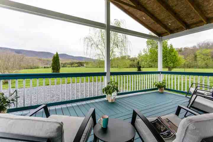 A covered deck features comfortable seating with a small table, offering a view of expansive green fields and distant mountains. Potted plants add a touch of nature, while the space is sheltered by a wooden roof, creating a relaxed outdoor atmosphere.