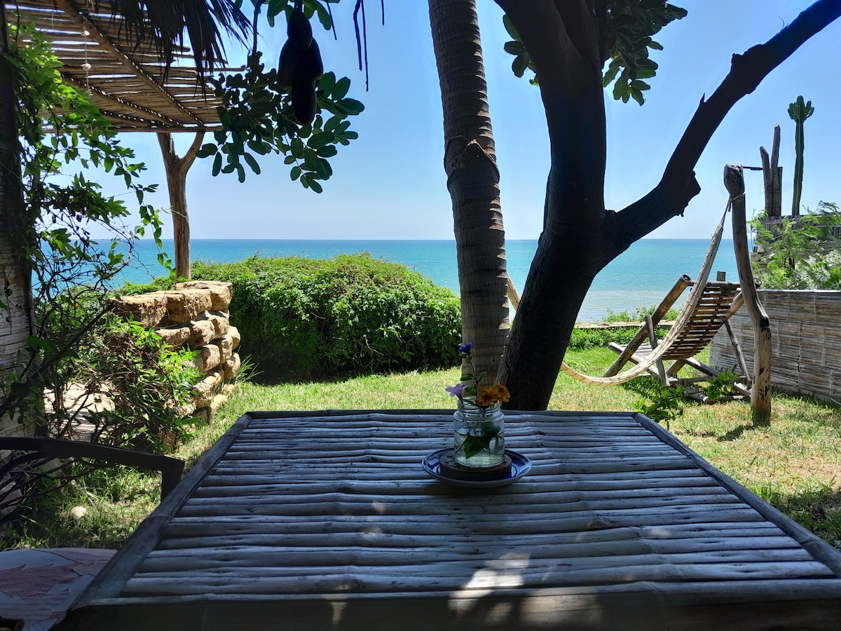 A table is set in a garden area, offering a serene view of the sea in the background. The table features a decorative vase with flowers. Nearby, a comfortable hammock and a lounge chair are positioned under the shade of palm trees, creating a relaxing outdoor space.