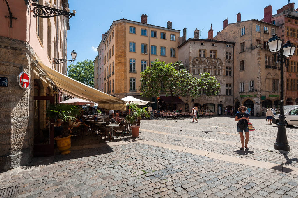 A bustling square is depicted with cafés featuring outdoor seating under umbrellas. Cobblestone pathways lead to historical buildings with vibrant architecture. Trees provide shade, and pedestrians are seen enjoying the lively atmosphere, while light filters through the clear blue sky.