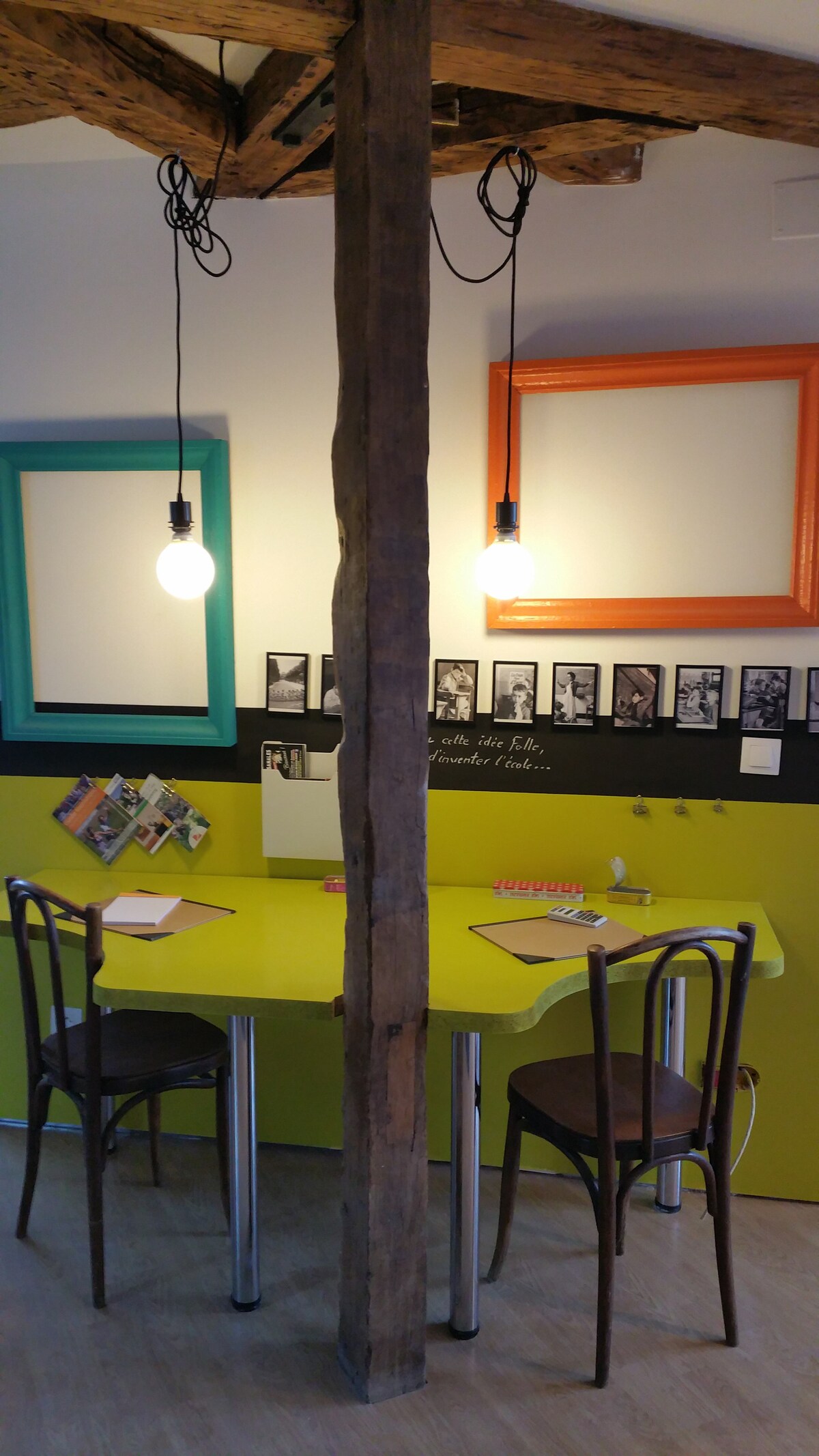 A vibrant dining area features a bright green table flanked by two chairs. The wall is adorned with framed artworks and photographs, alongside a section painted black for notes. Exposed wooden beams and pendant lights add warmth to the space.