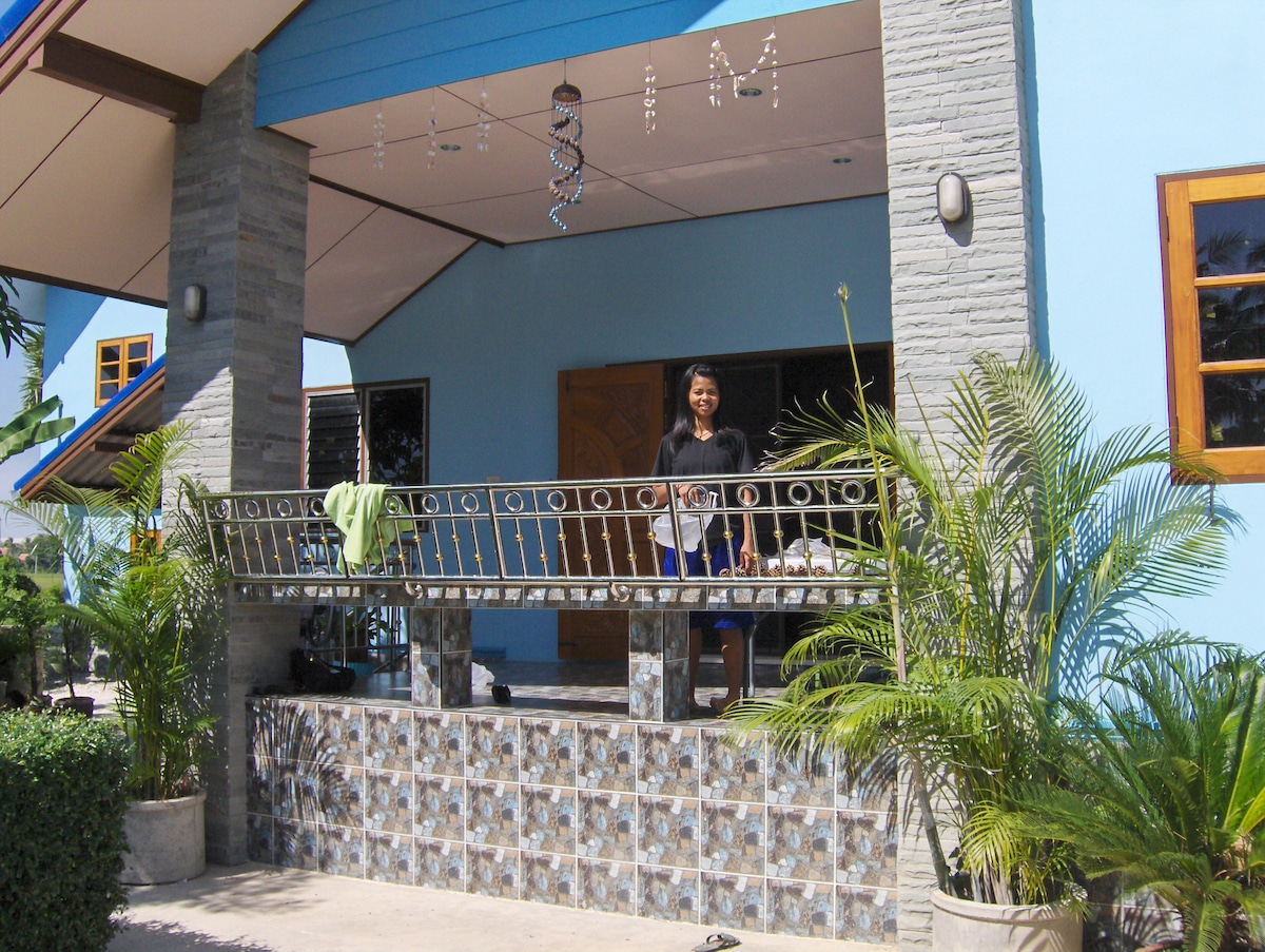 A covered porch features a decorative railing and is flanked by potted plants. A mosaic-patterned wall adds color to the setting. The double doors serve as an entrance to the home, and sunlight highlights the cool blue exterior and warm wooden window frames.