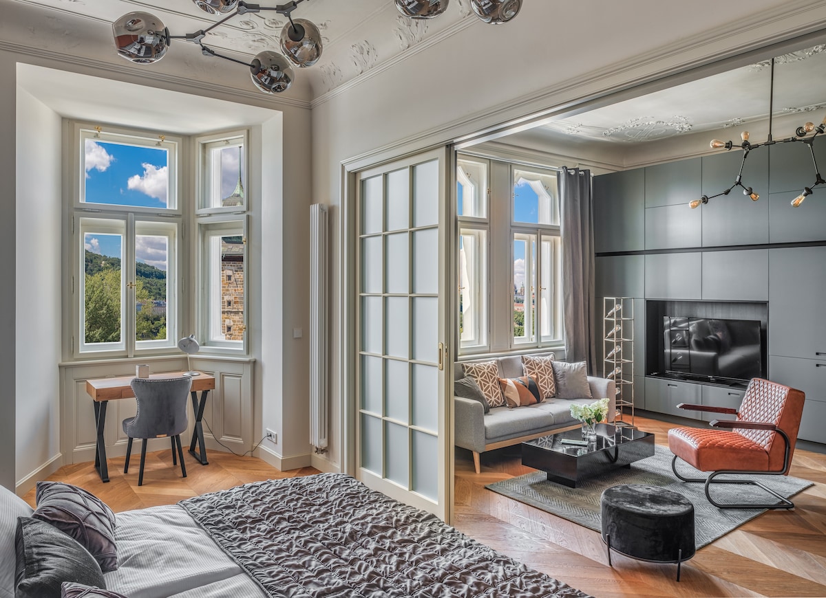 A spacious living area features large windows that provide natural light and scenic views. A comfortable seating arrangement includes a sofa and an accent chair, accompanied by a coffee table. A desk with a chair is positioned by the window, enhancing the inviting atmosphere.