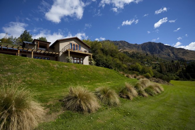 A well-maintained lawn leads to the two-story structure nestled on a gentle slope. The building is surrounded by a variety of shrubs and trees, with mountain peaks visible in the background under a bright blue sky dotted with clouds.