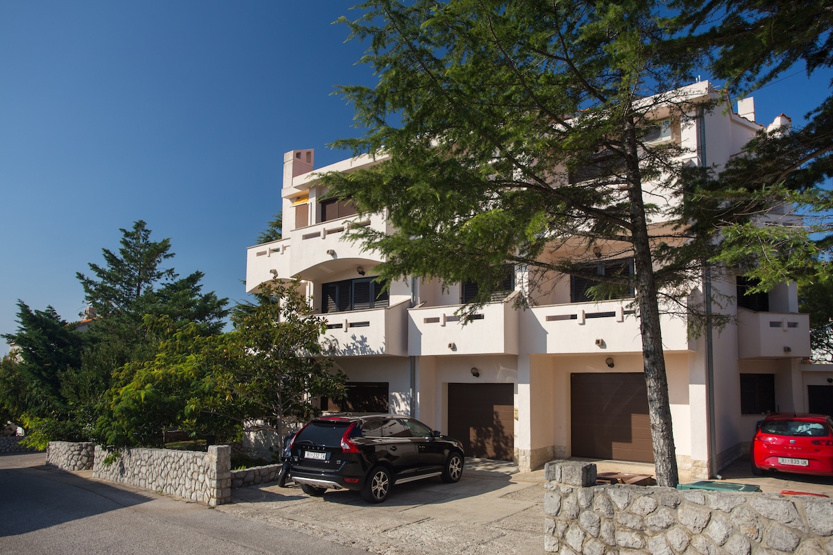 A three-story building with a light-colored exterior is flanked by tall trees and shrubbery. Two parked cars are visible in front of the building. The entrance area features large windows that allow natural light, contributing to the overall inviting appearance of the property.