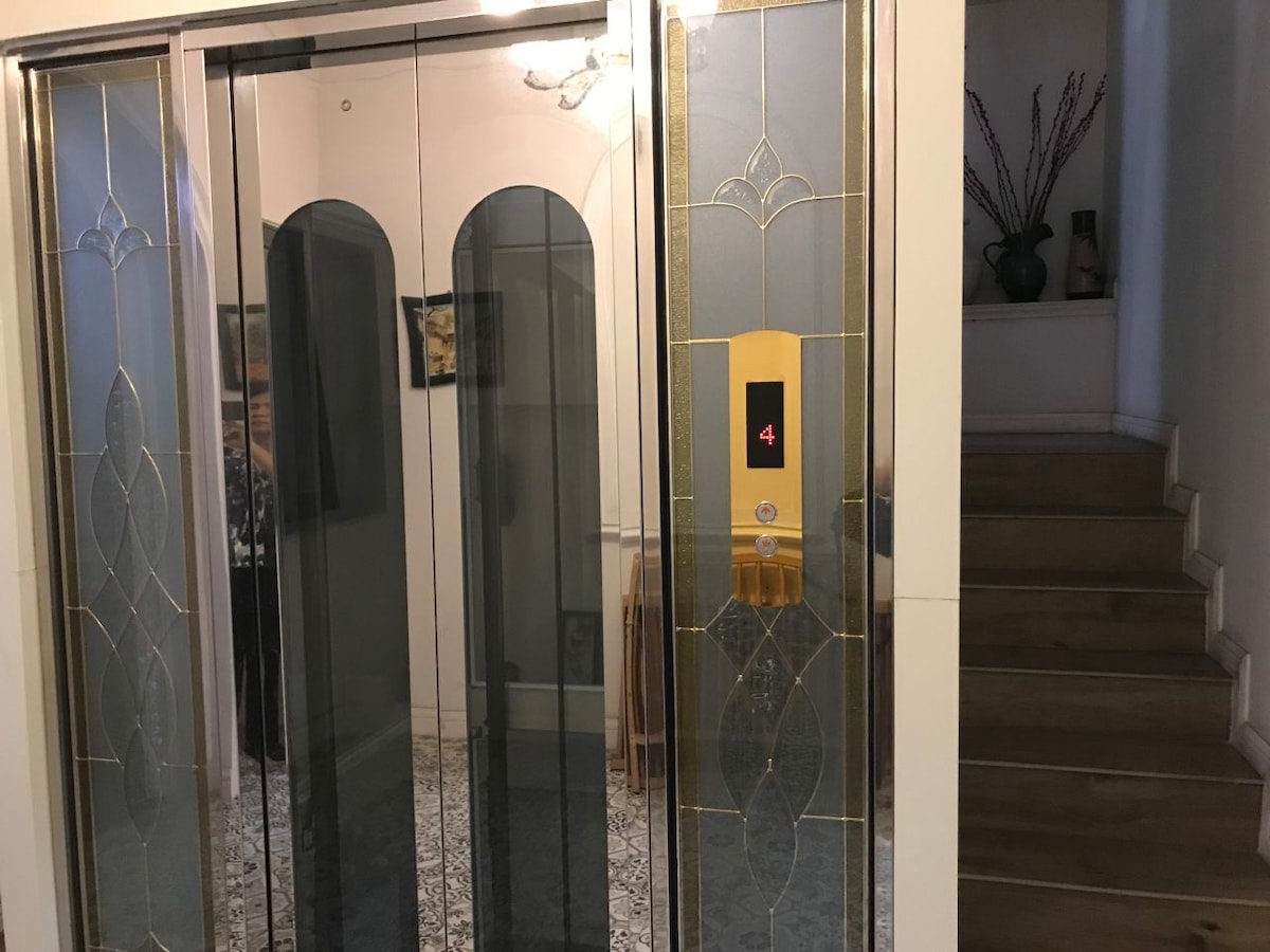 A modern elevator is shown with a decorative glass design and a digital display indicating the fourth floor. The surrounding area features a light-colored wall and staircase leading to an upper level, complemented by a decorative plant in a vase.