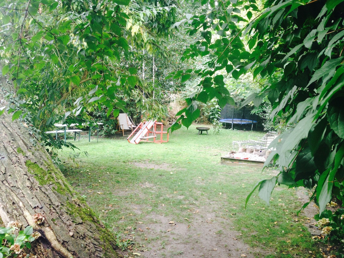 A lush garden space is surrounded by greenery, featuring a wooden play structure and a trampoline. A picnic table is positioned within the area, along with loungers set on a wooden deck. The ground is covered in grass, providing a natural setting.