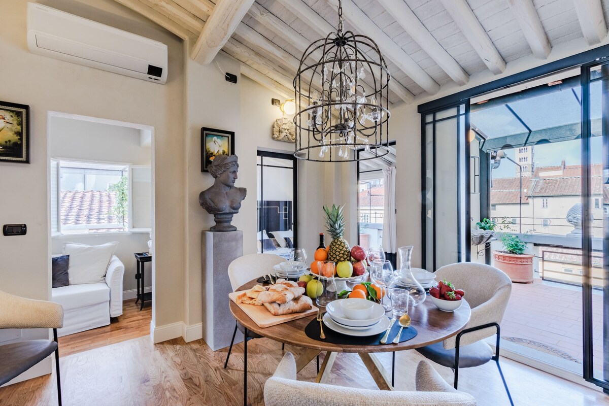 An elegantly furnished dining area is highlighted by a round wooden table set with plates and colorful fruits. A modern light fixture hangs above, complementing the airy space, while large glass doors provide access to the outdoor terrace and fill the room with natural light.