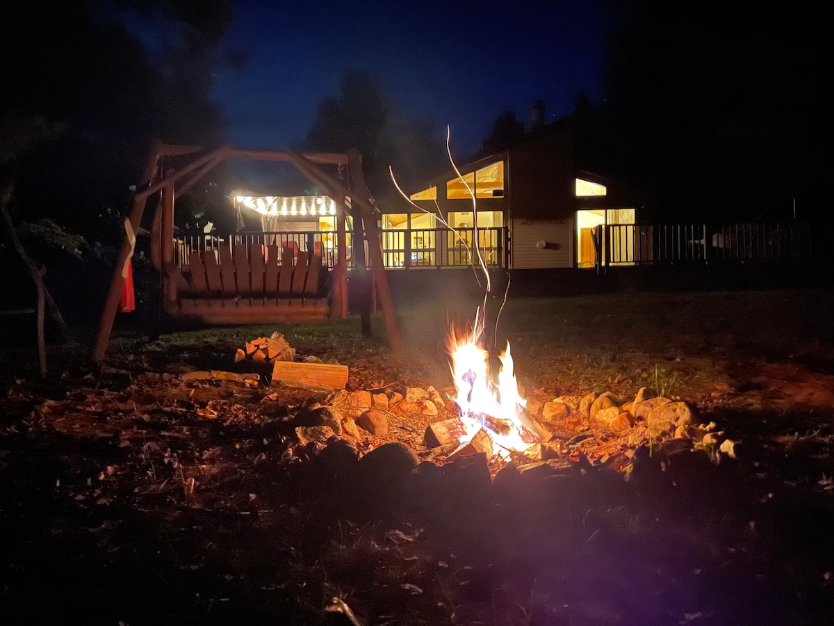 A fire pit surrounded by stones is illuminated as flames dance above the logs. A wooden swing is visible beside the fire, with warm light emanating from the cottage in the background, creating a cozy evening setting.