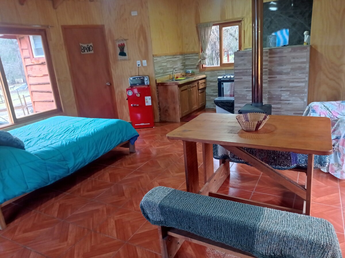 An inviting interior features a spacious bed covered with a blue quilt. A wooden table with benches is positioned in the center, while a small kitchenette is visible in the background. Natural light enters through the window, highlighting the warm wood paneling.