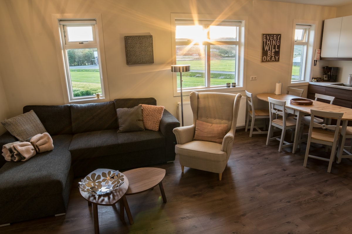 A bright living area is depicted with a dark gray sofa and a light-colored armchair. Natural light enters through the windows, highlighting a round coffee table and a dining table with chairs. A motivational wall decor is visible, enhancing the cozy ambiance.