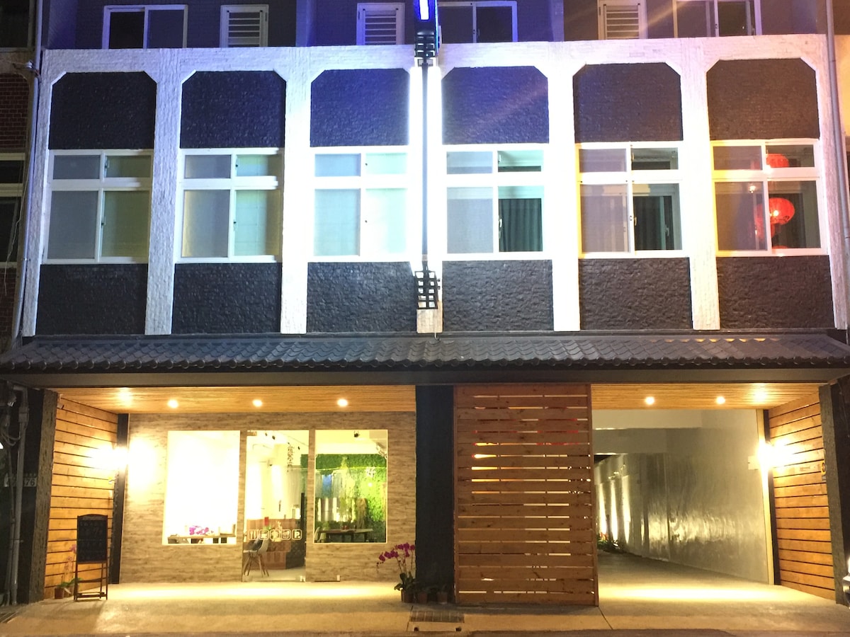 The building presents a modern façade with a combination of dark and light elements. Large windows provide ample visibility into the interior space. Warm lighting enhances the welcoming atmosphere at night, while a wooden entrance creates a subtle contrast with the exterior.