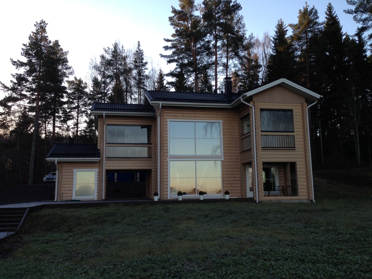 A modern log cabin is presented, featuring large glass windows that allow natural light to fill the interior. The structure is surrounded by trees, with a clear view of the private outdoor space. Multiple balconies enhance its architectural appeal.