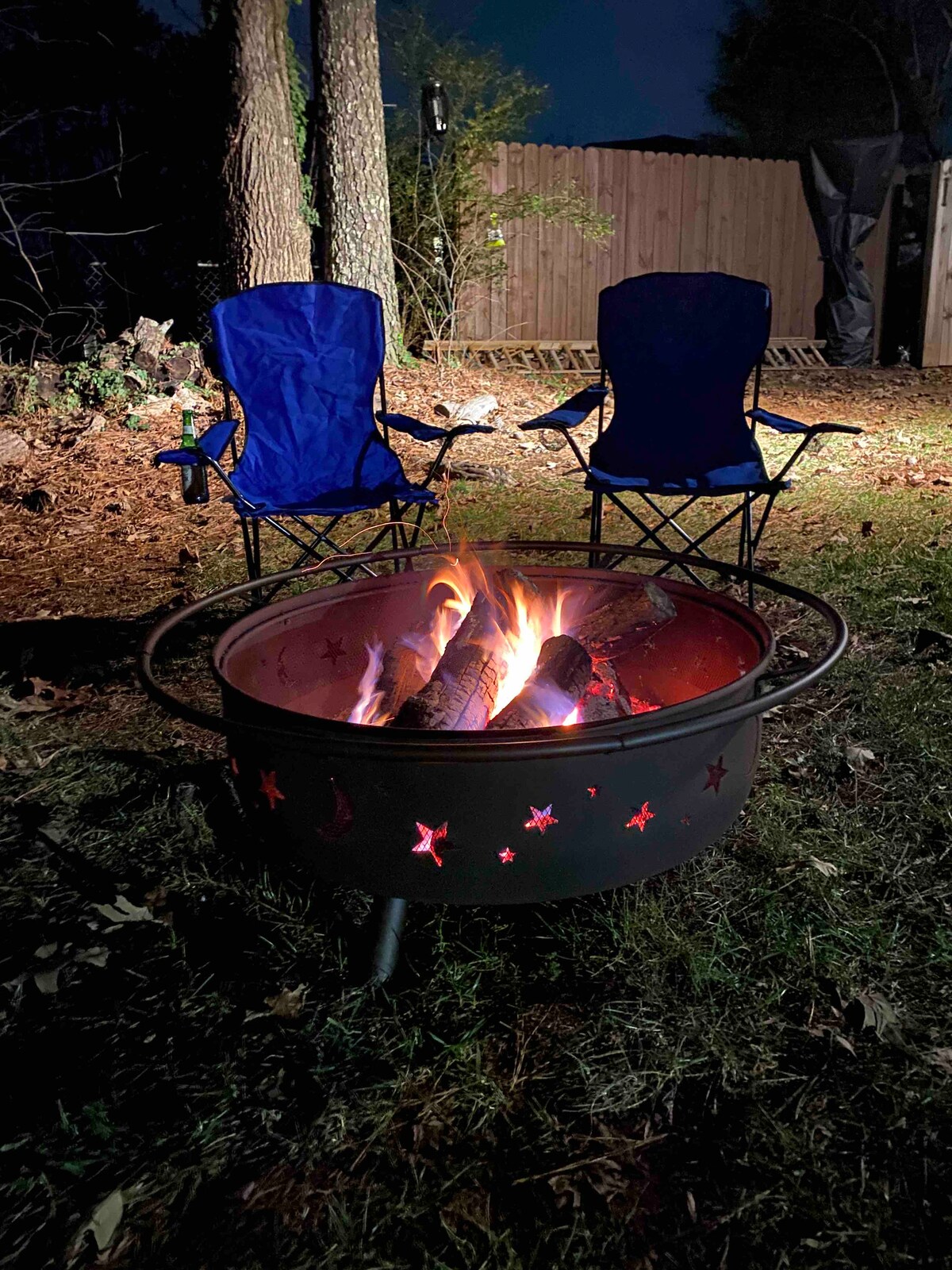 A fire pit featuring a flame surrounded by logs is set in a grassy area. Two blue camping chairs are placed nearby, positioned for relaxation. Surrounding trees can be seen in the background, creating a serene outdoor space, especially suitable for evening gatherings.