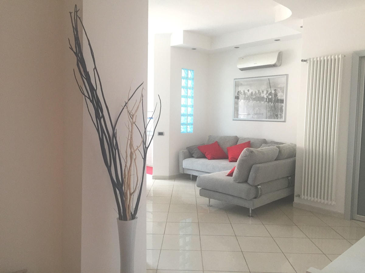 A contemporary living area is presented with a light-colored sectional sofa featuring red accent pillows. A tall vase with dried branches adds a decorative touch. A large photograph on the wall complements the soft lighting and glossy tiled floor, enhancing the open ambiance.