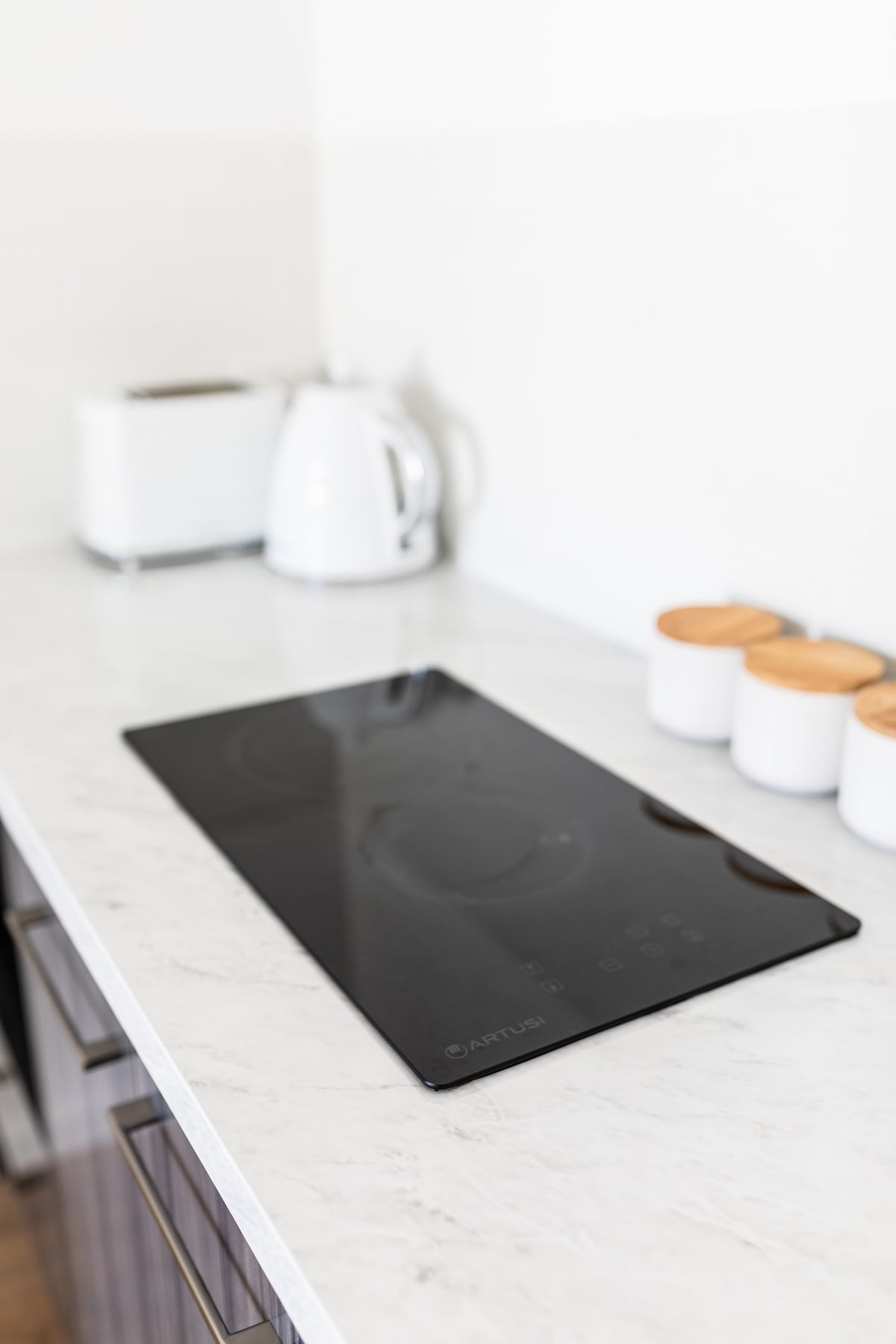 A sleek black induction cooktop is positioned on a white marble countertop. Behind, a silver kettle and a white toaster are visible. Three white containers with wooden lids sit neatly on the counter, adding a touch of warmth to the modern kitchen setup.