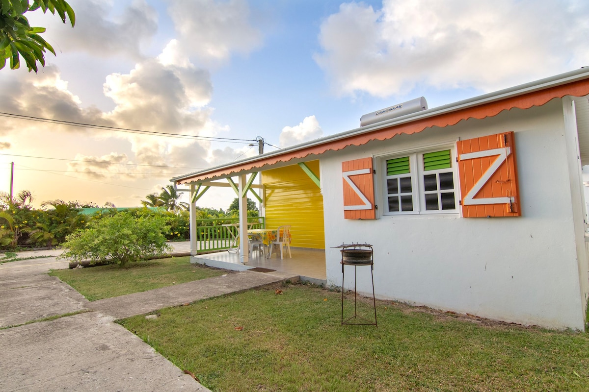 A charming exterior is displayed with a light-colored cottage featuring colorful orange shutters and green accents. A covered porch area includes a small table and chairs. The surrounding lawn is well-maintained, complemented by a bright sky with scattered clouds.