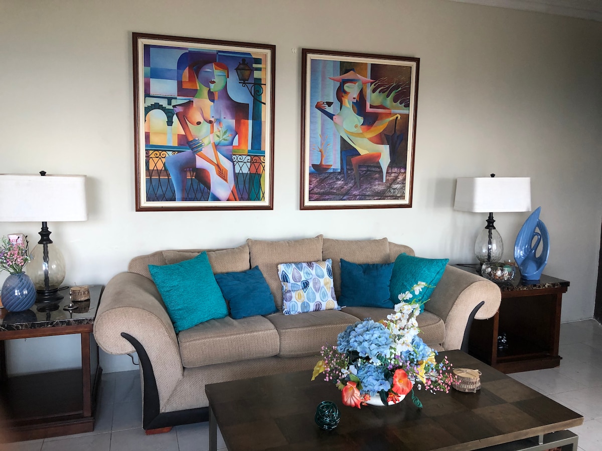 A spacious living area features a comfortable beige sofa adorned with turquoise and patterned throw pillows. Two vibrant artworks hang on the wall above the sofa. A coffee table with floral arrangements sits centrally, complemented by decorative lamps on either side.
