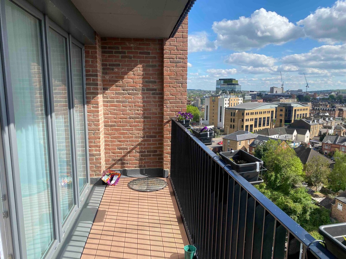 A modern balcony is featured, showcasing a brick wall and expansive city views. Potted plants are arranged along the railing, and patterned outdoor decor is positioned on the tiled floor. Natural light illuminates the space, enhancing the open-air atmosphere.