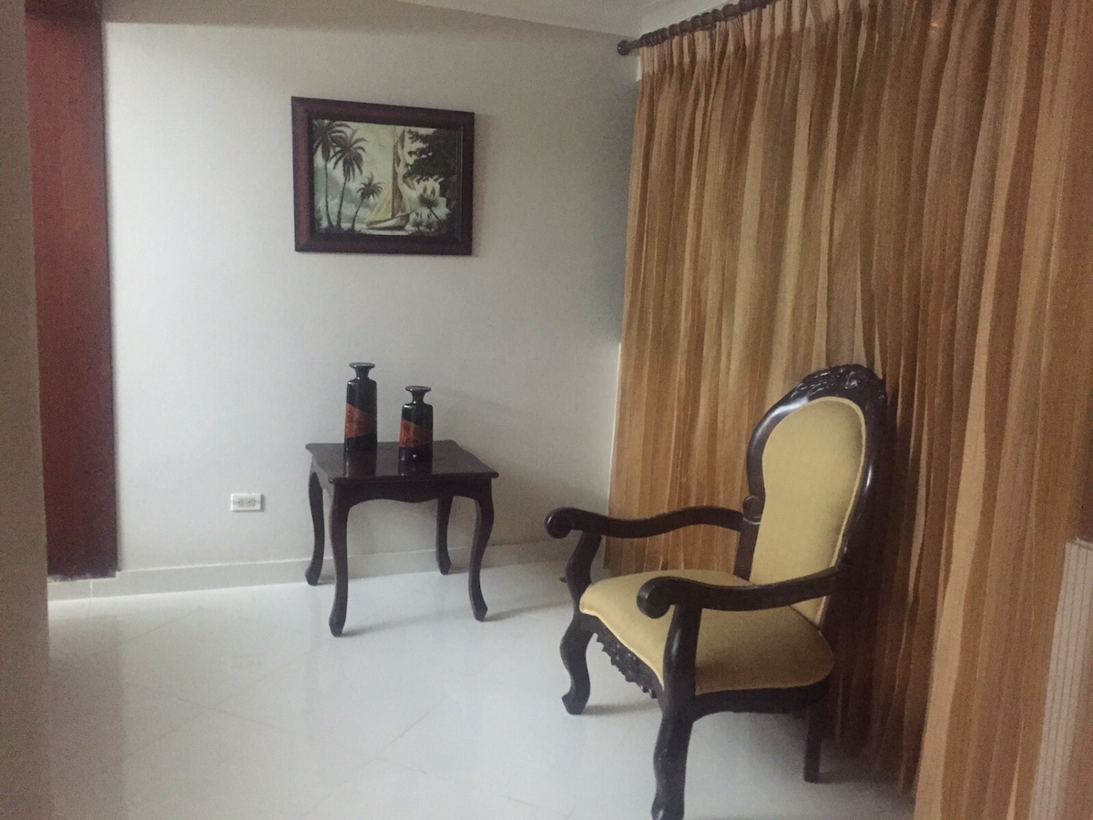A tastefully furnished corner features a yellow armchair next to a dark wood table. Two decorative vases are placed on the table, with a wall art piece depicting palm trees hanging above. Light beige curtains frame a window, creating a serene ambiance.