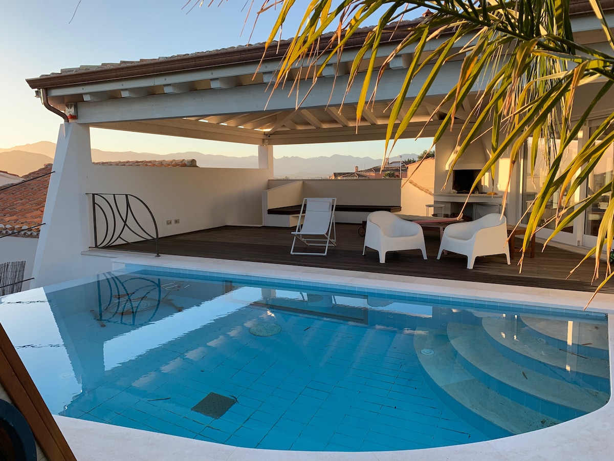 A sunlit terrace features a private swimming pool, reflecting the surrounding mountain landscape. Comfortable lounge chairs are arranged nearby, complemented by a shaded seating area under a stylish canopy. The serene atmosphere is enhanced by the tranquil water and clear sky.