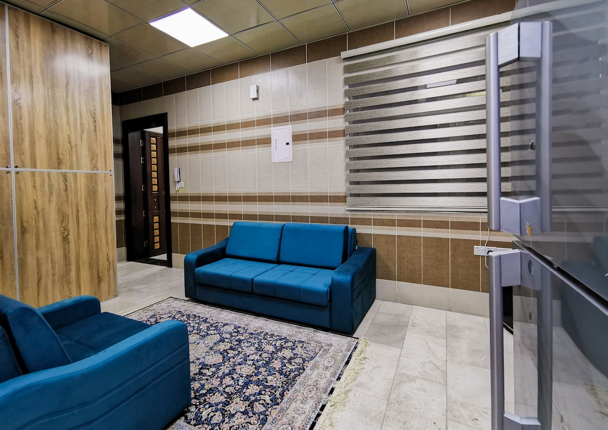 A modern living area features two teal sofas set on a patterned area rug. Natural light enters through horizontal blinds, illuminating the stylish decor. The walls are adorned with neutral tiles, and a wood-paneled partition adds texture to the space.