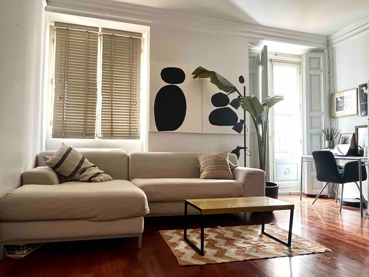 A bright living area features a light-colored sectional sofa with decorative pillows, positioned beside a coffee table. A large piece of abstract art hangs on the wall, complemented by a tall potted plant. Natural light filters through the window with wooden shutters.