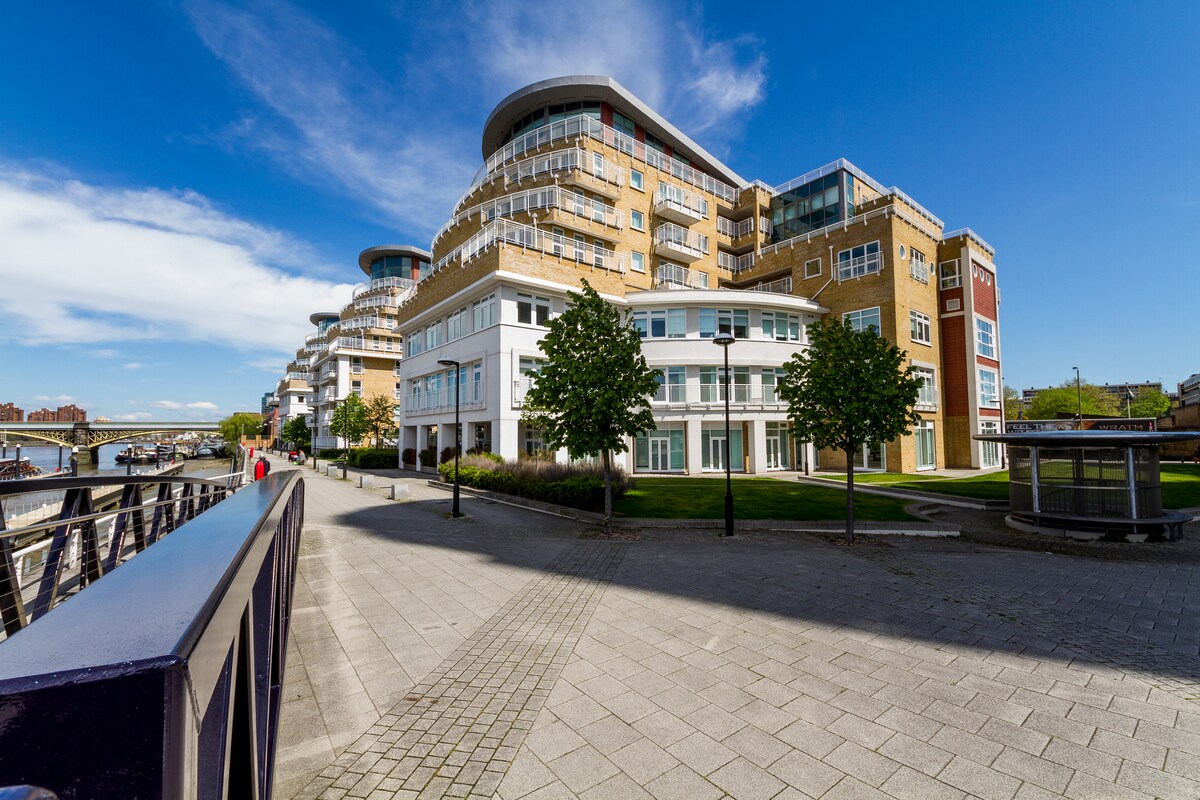 A modern building complex is shown, featuring multiple stories with rounded balconies. The structure is complemented by lush greenery and well-maintained walkways. Sunlight enhances the brick facade, while a nearby riverbank is visible in the background, promoting a serene outdoor space.