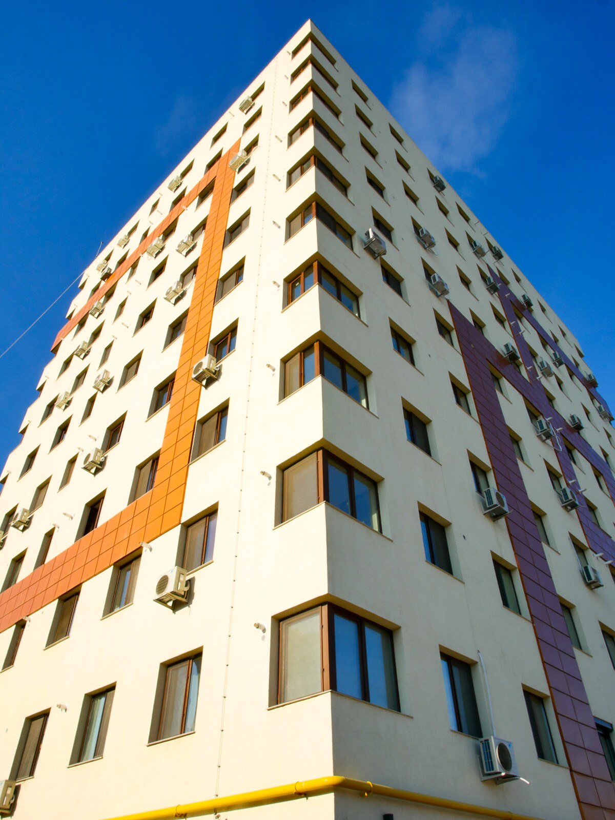 The residential complex is a modern, multi-story building featuring a textured facade with varied color panels. Multiple air-conditioning units are mounted on the exterior. Windows are arranged symmetrically, allowing for ample natural light.