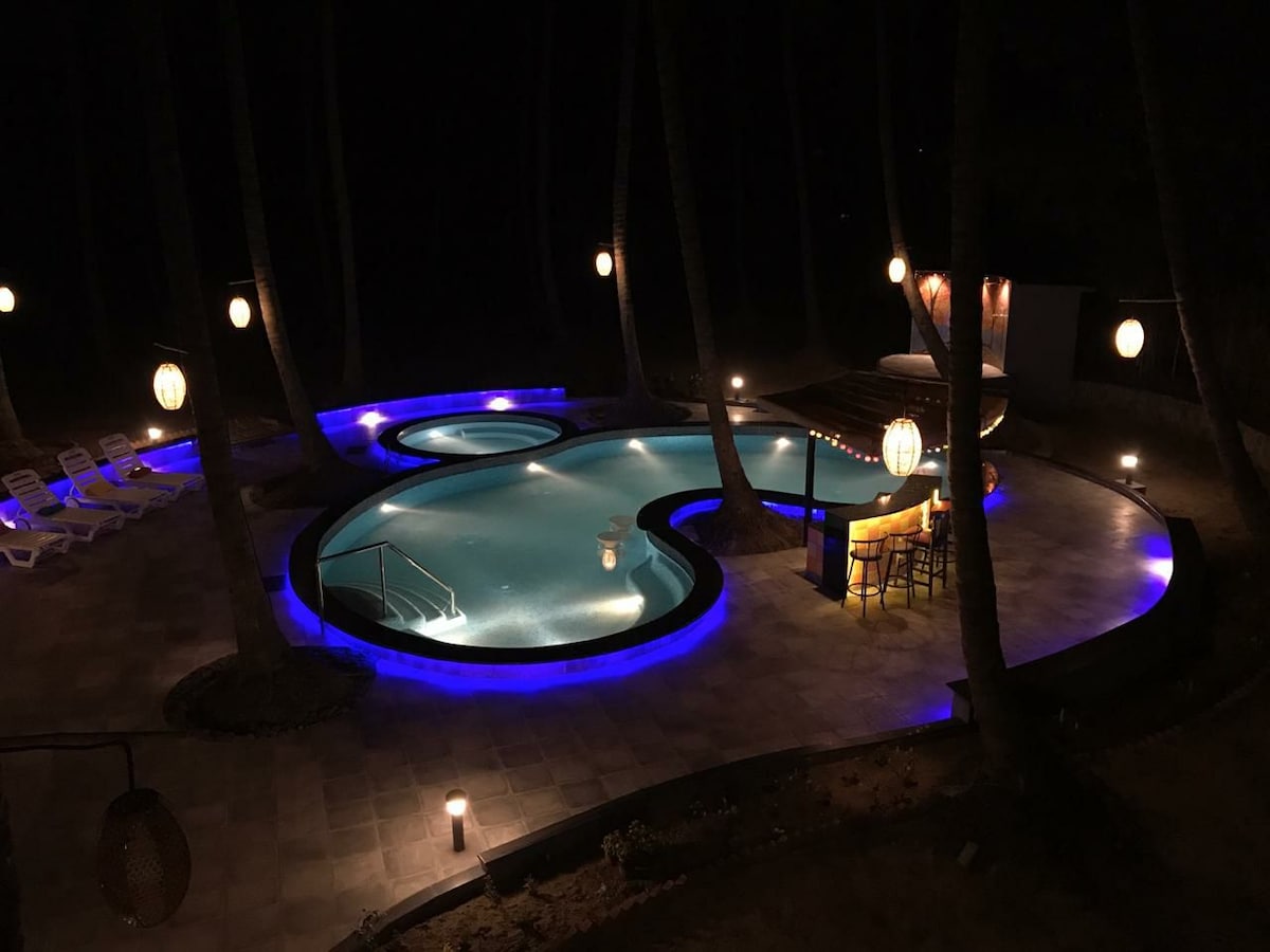 A uniquely shaped pool is illuminated by blue lighting, surrounded by palm trees and lounge chairs. Soft light fixtures hang from overhead, casting a gentle glow over the area. A seating arrangement lies near the pool, providing a relaxing outdoor space.