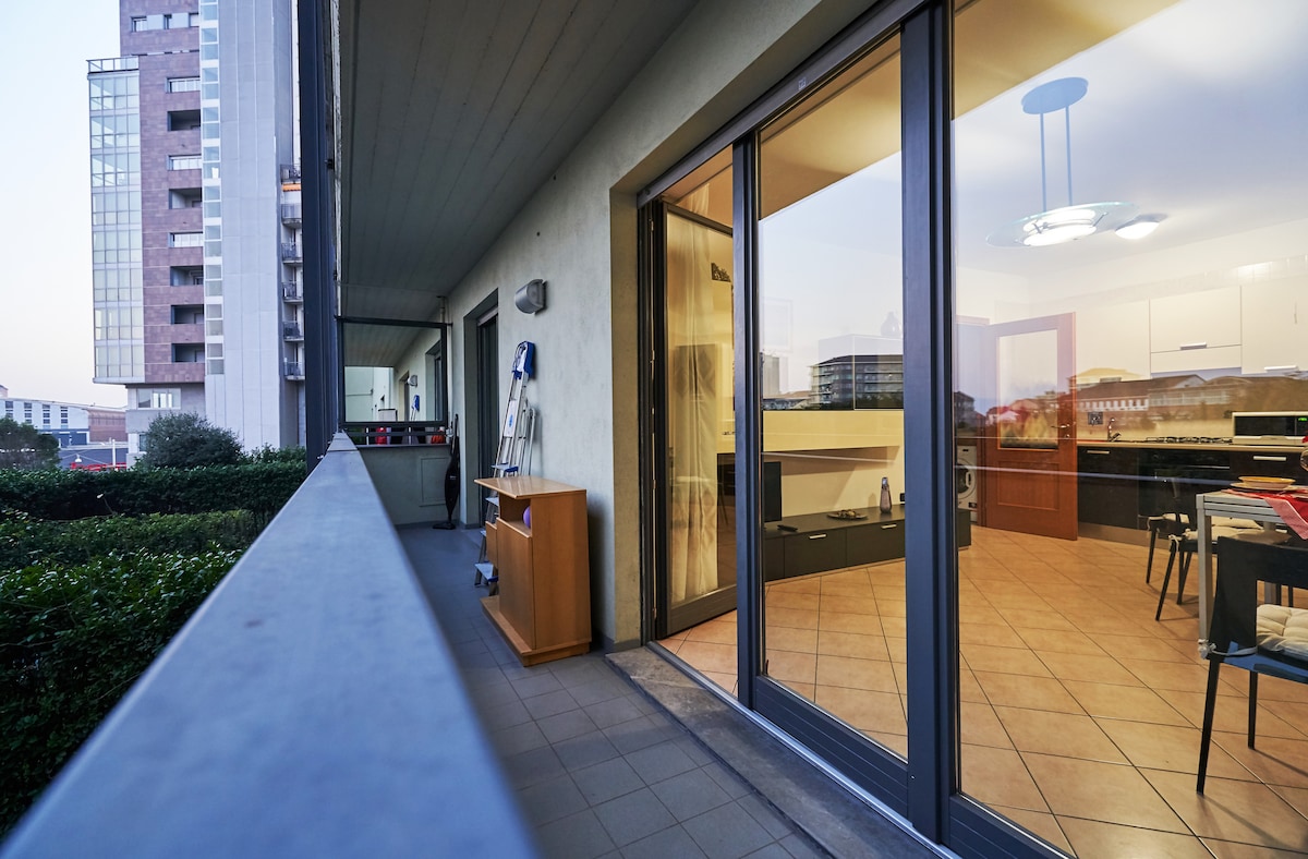 A wide balcony overlooks a well-lit kitchen area through large glass doors. A wooden cabinet is positioned against the wall, while the tiled flooring is visible. Soft evening light enhances the welcoming atmosphere of the space.