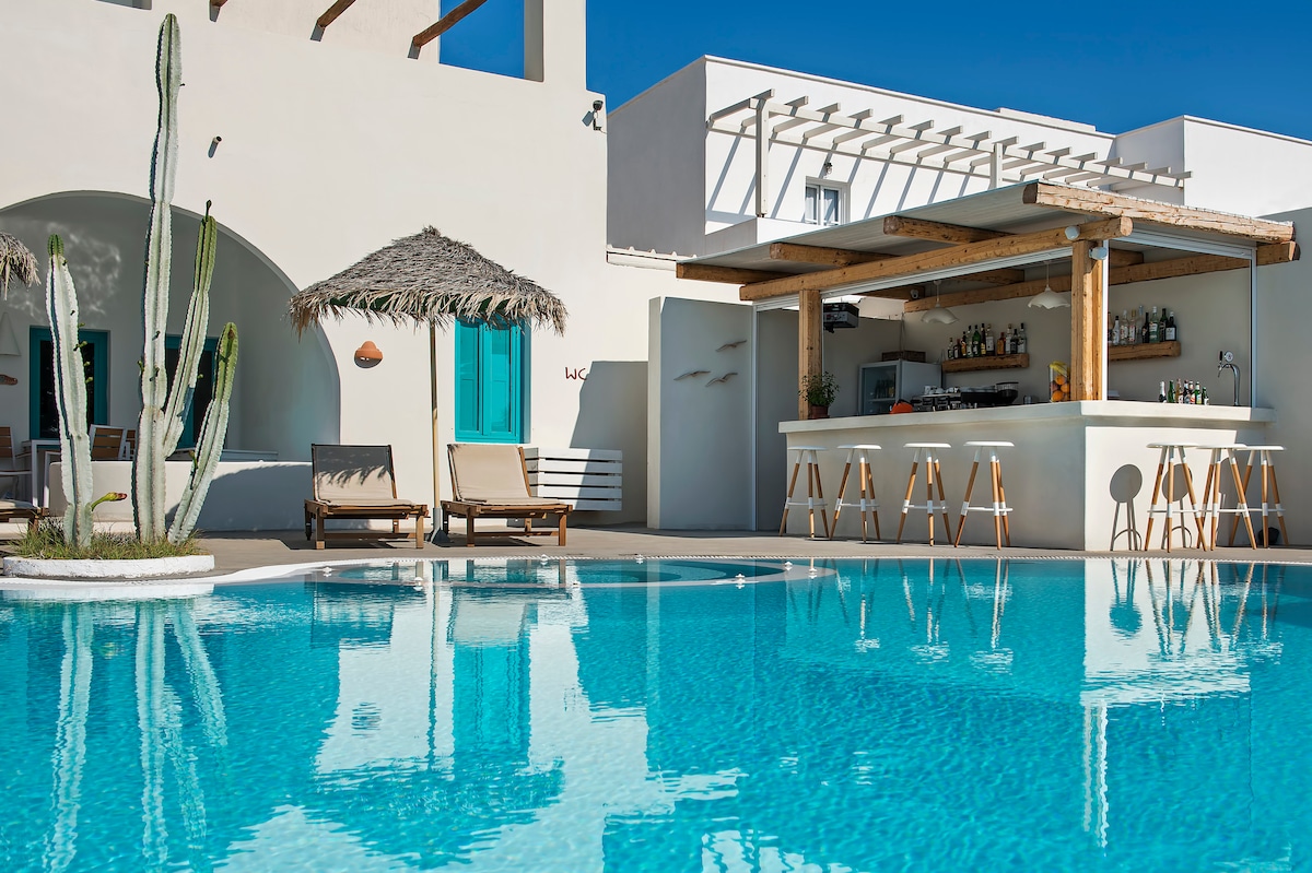A clear turquoise pool reflects the sunny sky and surrounding architecture. A shaded bar area with high stools is positioned near the water. Lounge chairs are neatly arranged beside the pool, offering a space for relaxation amidst well-maintained greenery.
