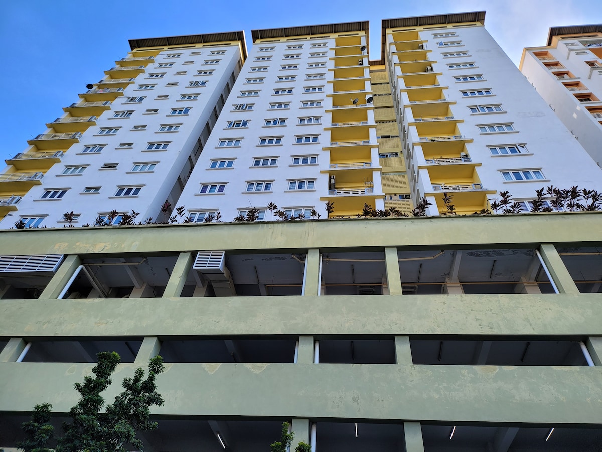 A tall residential building is depicted, showcasing multiple levels with a mix of white and yellow exterior. The ground floor features a parking area, while greenery is visible on the upper balconies. The blue sky serves as a backdrop, enhancing the building's structure.