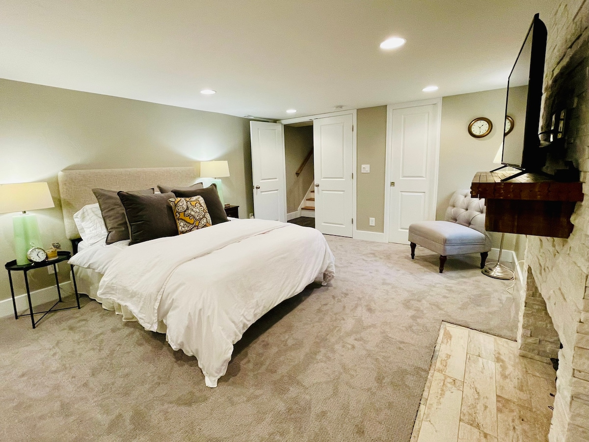 A spacious bedroom is presented with a queen bed dressed in white linens and accented with decorative pillows. A soft gray armchair complements the room, which includes a mounted television and a fireplace. Natural light fills the space through recessed lighting.