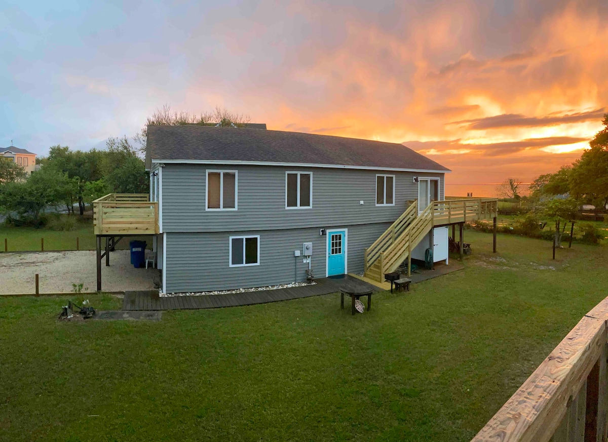 The exterior of a two-story house is captured during sunset, highlighting its gray siding and distinctive blue door. Wood steps lead to the upper deck, surrounded by a grassy yard and trees. The colorful sky adds warmth to the scene, emphasizing the tranquil setting.