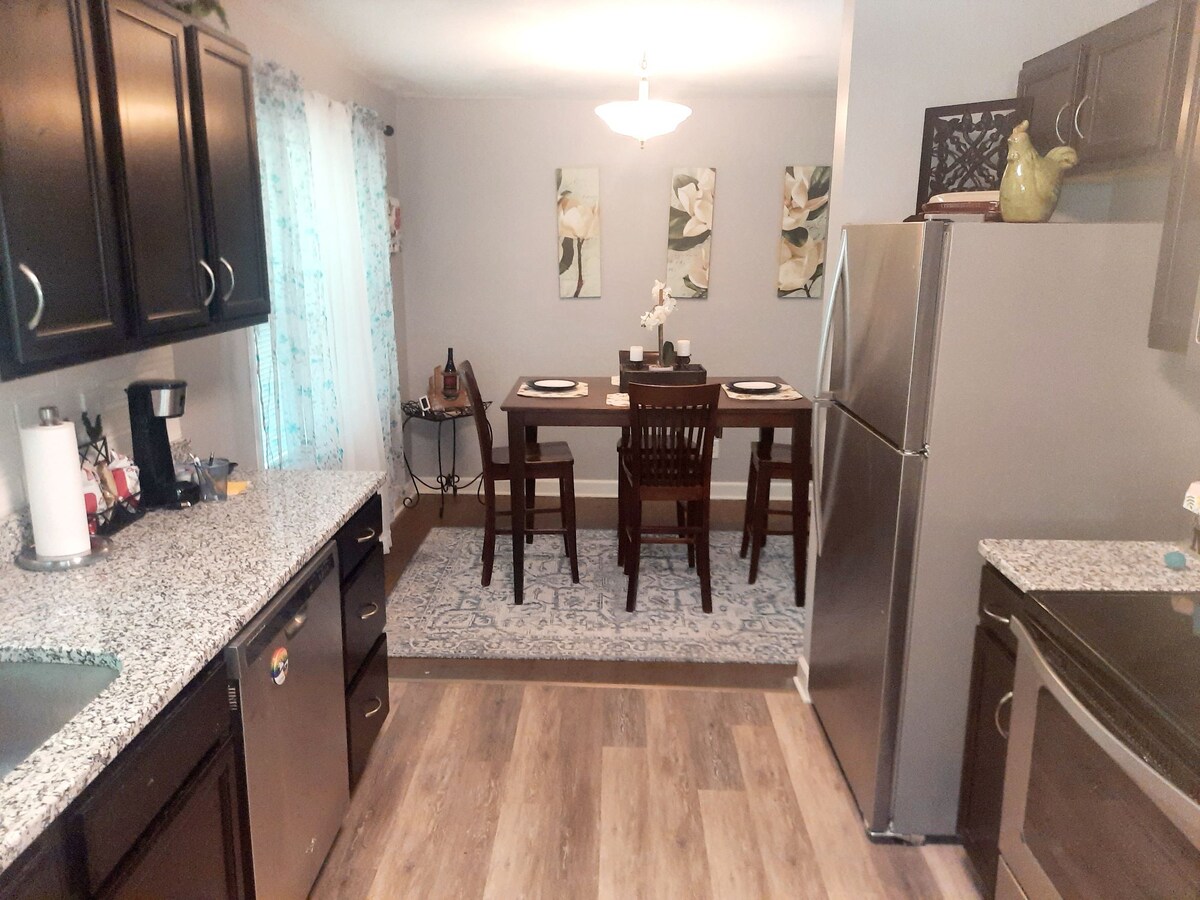 The kitchen area features dark cabinetry and a granite countertop, with silver appliances visible. A dining table with four chairs is set in the adjoining space, accompanied by decorative wall art and a light fixture overhead. Soft natural light filters through sheer curtains.