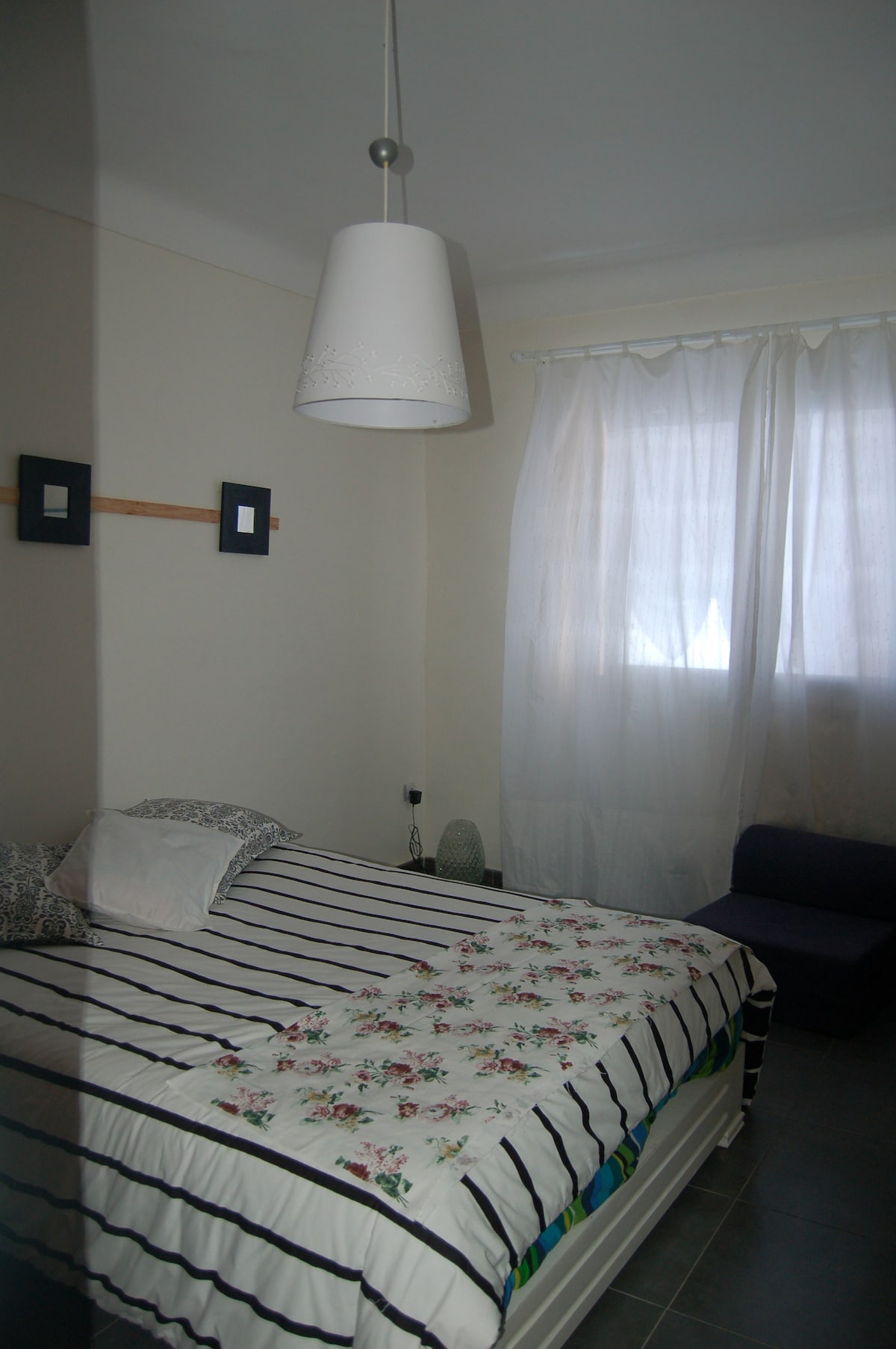 A cozy bedroom features a spacious bed with a striped duvet and floral accents. A window is dressed with sheer white curtains, allowing natural light to illuminate the room. A small seating area is provided by a dark-colored couch against the wall.