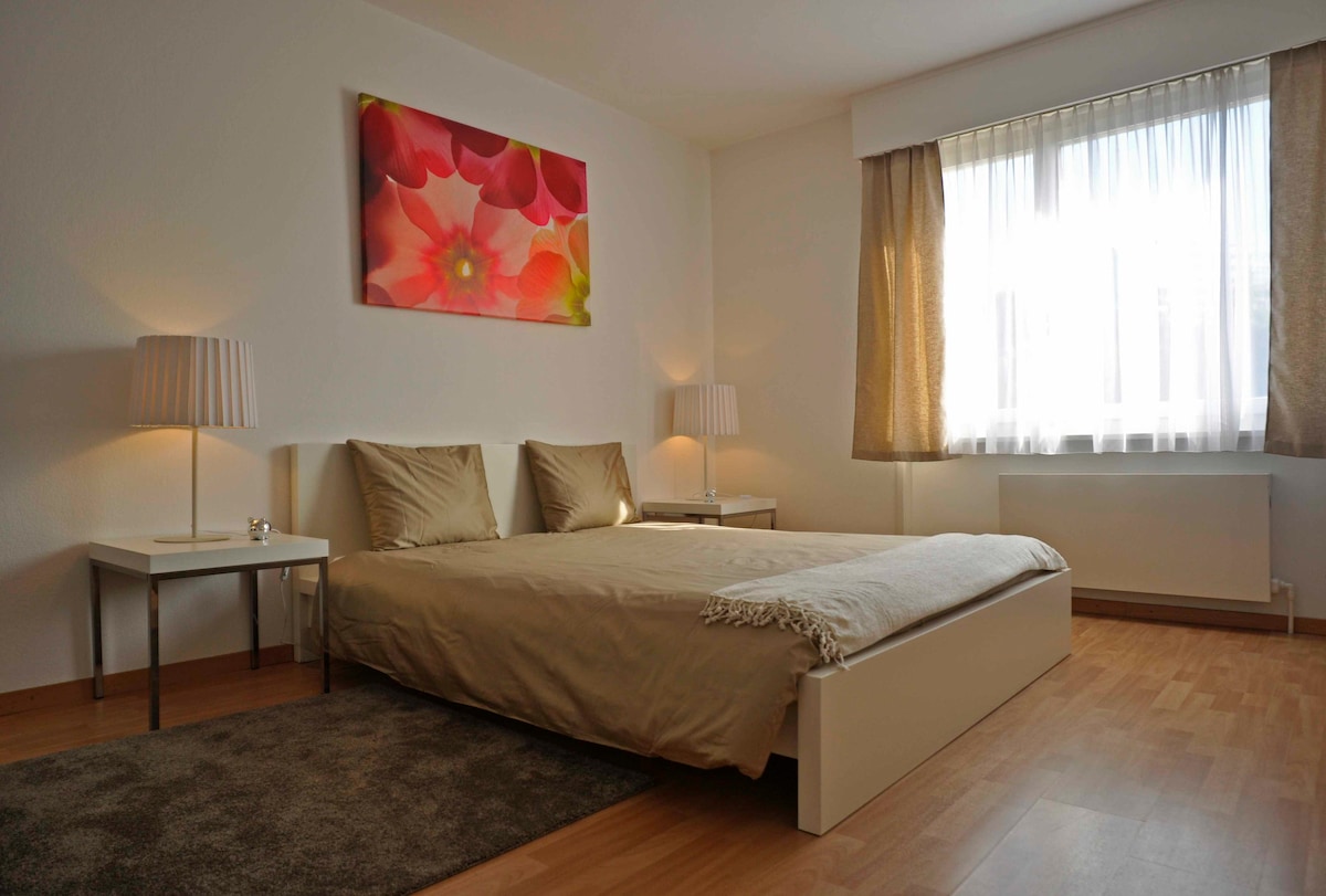 A spacious bedroom features a light-colored bed with soft bedding, complemented by two bedside tables with lamps. A colorful floral artwork decorates the wall above the bed. Natural light filters through a large window, adorned with light drapes, enhancing the serene atmosphere.