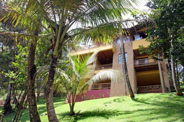 A multi-level house is nestled among lush green palms and tropical foliage. The structure features multiple balconies with wooden railings, providing an inviting connection to the surrounding nature, while a well-maintained lawn leads up to the entrance.