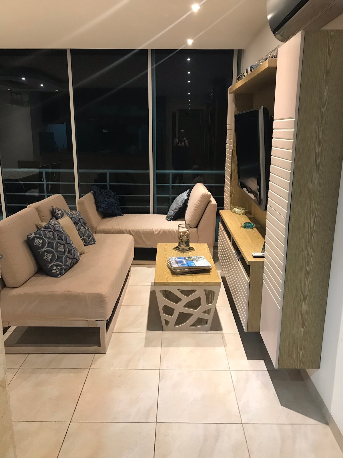 The living area features a light-colored sofa adorned with decorative pillows, positioned next to a wooden coffee table. A television is mounted on the wall opposite large glass windows that allow for ample natural light, illuminating the space.
