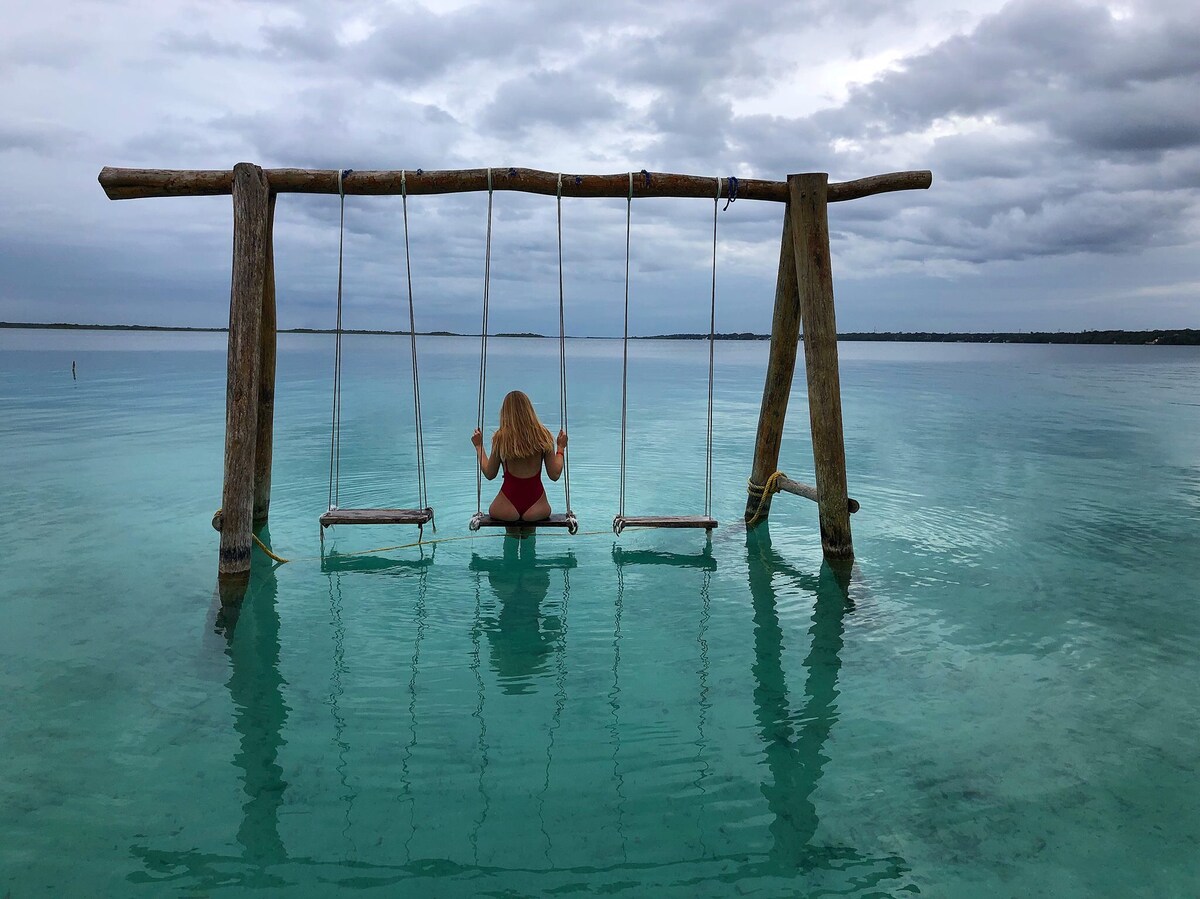 A wooden swing is positioned over calm turquoise waters, allowing for relaxation and play. The sky appears cloudy, creating a serene atmosphere. The swing's sturdy frame is crafted from rough wood, while the gentle waves reflect the soft tones of the environment.