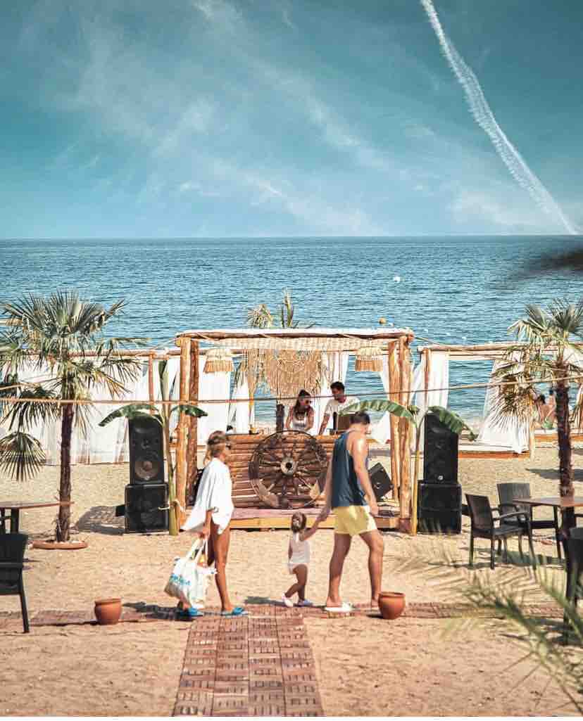 A scenic beachfront setup features a wooden stage with musicians, surrounded by palm trees. Sun loungers and chairs are arranged in the sandy area. Calm waters are visible in the background, with a clear blue sky above. A family walks along the beach, enjoying the atmosphere.