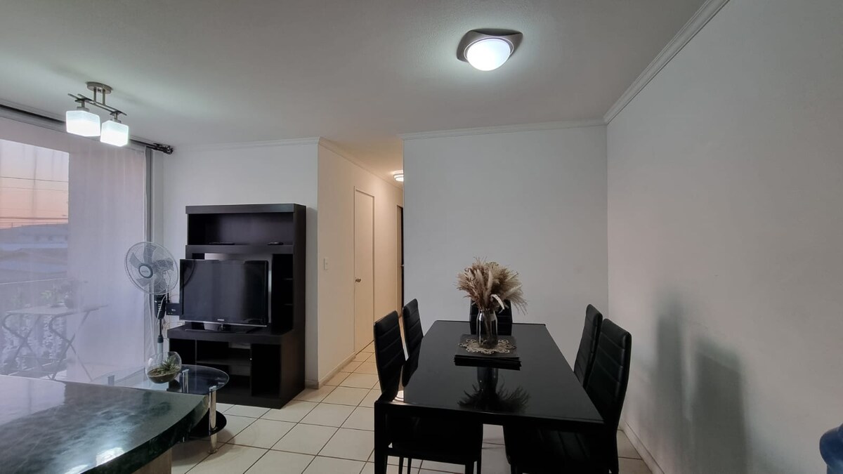 A dining area is furnished with a dark table and six chairs, set against a backdrop of a sleek black entertainment unit. A fan is placed nearby, and soft natural light enters through a window with sheer curtains.