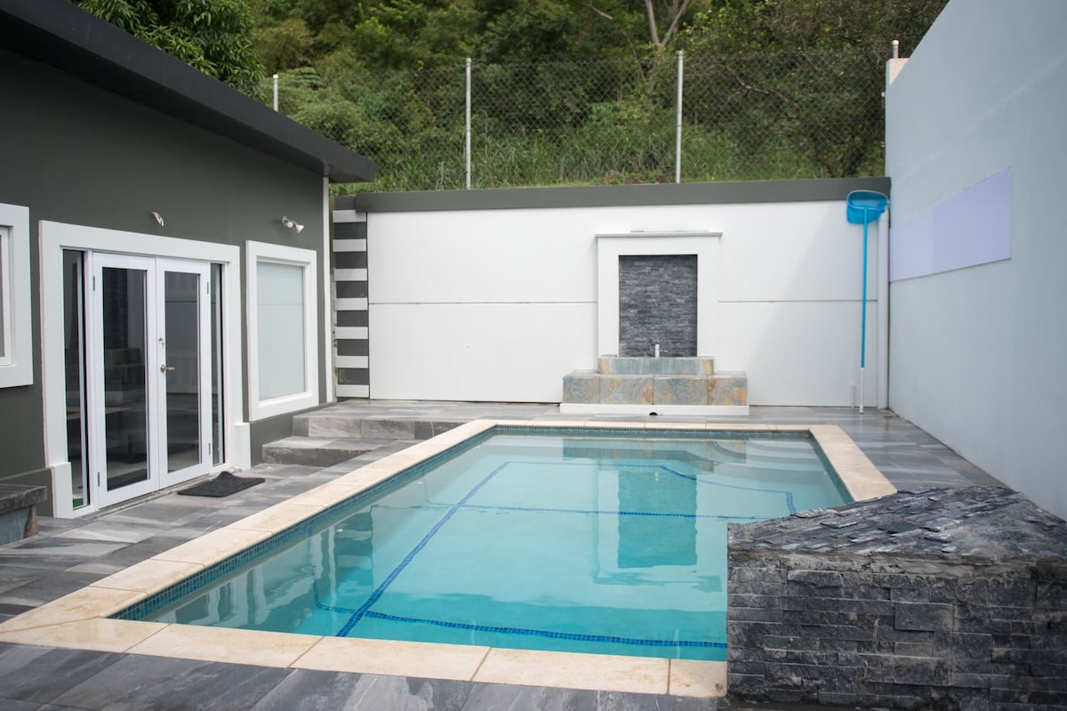 A serene outdoor area features a clear swimming pool bordered by sleek gray tiles. Surrounding walls are finished with smooth, light-colored surfaces, enhancing the open feel. Natural elements are visible in the background, providing a tranquil backdrop.