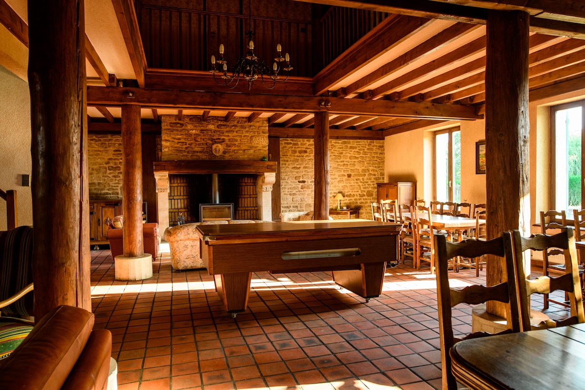 A spacious communal area features a large billiard table in the center, surrounded by wooden posts and a cozy seating arrangement. Natural light floods through large windows, highlighting the rustic stone walls and wooden beams that create a welcoming atmosphere.