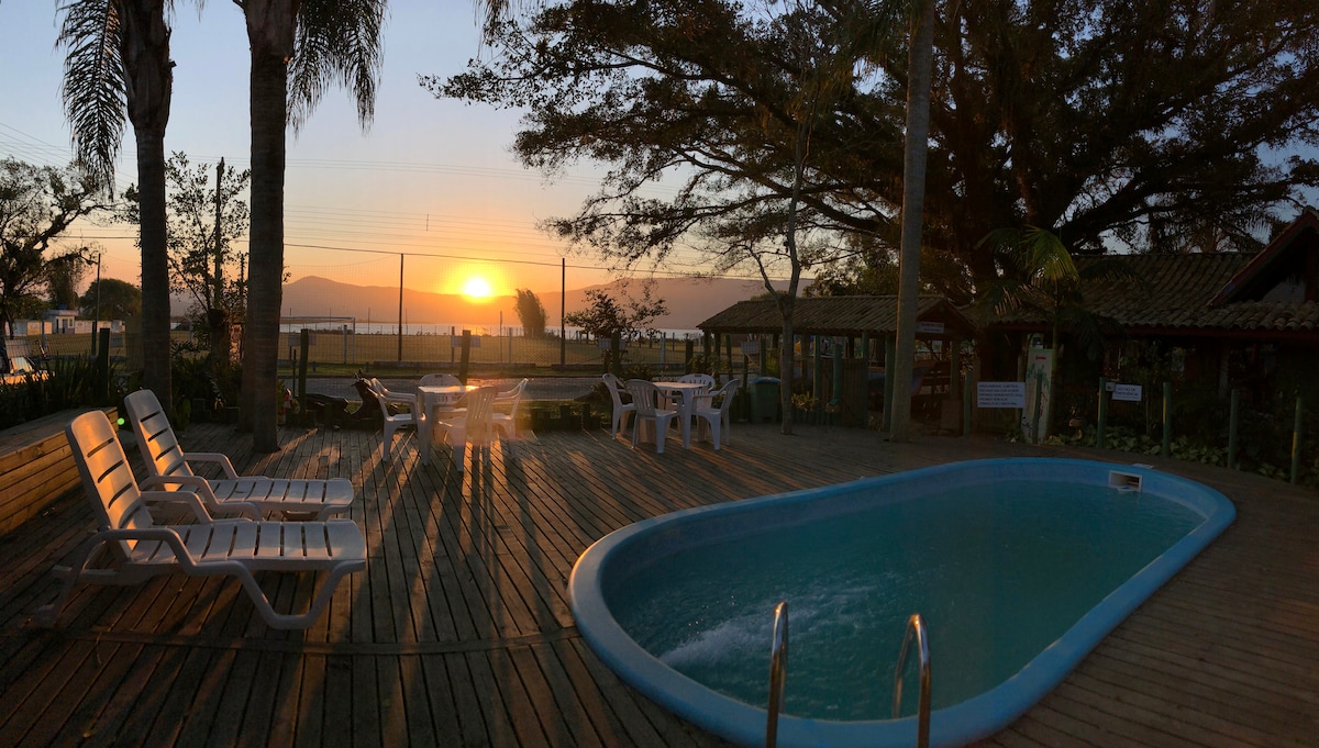 A poolside area is shown with a swimming pool surrounded by wooden decking. Sun loungers are positioned nearby, offering a view of the sunset over the horizon. Lush trees provide shade and a sense of tranquility to the outdoor space.