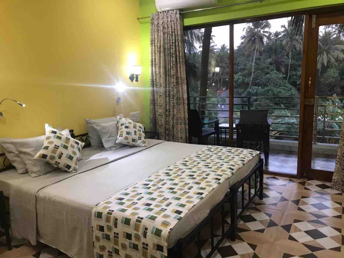 A well-lit room features two spacious beds with light-colored linens and a patterned throw blanket. Floor-to-ceiling windows offer views of surrounding greenery. A small dining table and chairs are positioned near the door leading to the balcony.