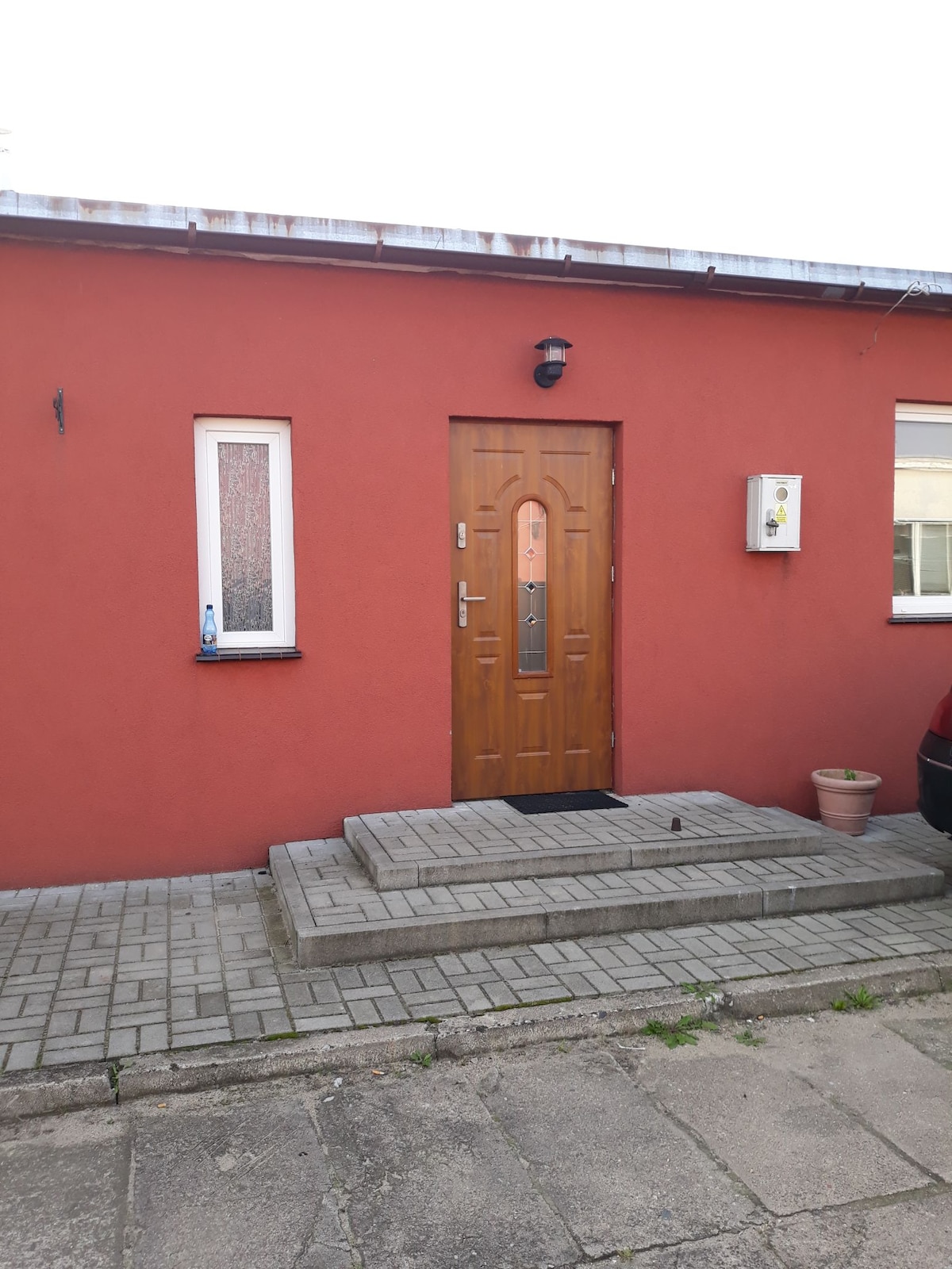 A welcoming exterior features a warm red facade with a wooden door framed by a small rectangular window on the side. Steps lead to the entrance, surrounded by paved flooring. A single flower pot is positioned near the wall, adding a simple touch to the entrance.