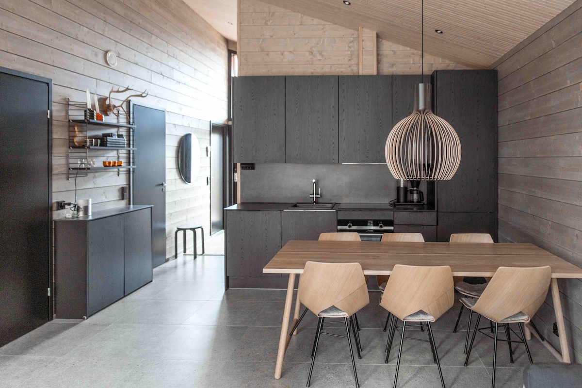 A modern kitchen and dining area features sleek black cabinetry and a spacious wooden dining table surrounded by six light-toned chairs. A distinctive pendant light hangs above the table, complementing the natural wood walls and creating a harmonious space for dining and cooking.